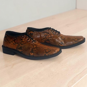 May include: A pair of lace-up oxford shoes featuring a brown and black snakeskin-textured pattern. The shoes have a low-profile silhouette, a rounded toe box, black laces, and a solid black sole that provides a distinct contrast to the patterned upper.