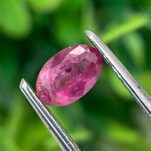 May include: A faceted oval-shaped pink gemstone held by silver-toned metal tweezers. The stone displays a vibrant magenta hue with internal clarity and light-reflecting facets across its surface, showcasing a polished and transparent appearance.