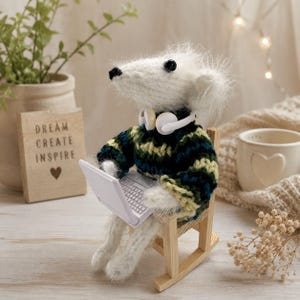 May include: A fuzzy white crocheted mouse figurine wearing a dark green and yellow striped jumper and white headphones. The mouse sits on a small wooden rocking chair, holding a miniature white laptop computer on its lap.