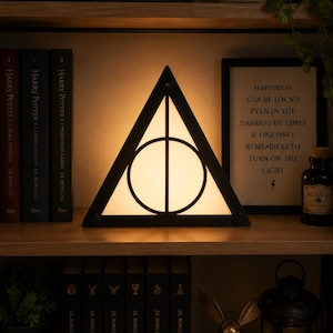 Puede incluir: Una lámpara triangular con el símbolo de las Reliquias de la Muerte en negro, con una luz cálida y brillante que emana desde detrás del marco. El diseño consta de un triángulo, un círculo y una línea vertical con retroiluminación.