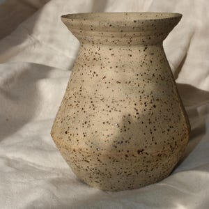 Könnte beinhalten: Beige Keramikvase mit ausgestelltem Rand und gesprenkelter, strukturierter Oberfläche. Die Vase hat einen leicht konischen Körper und ist mit dunkelbraunen Sprenkeln bedeckt. Das Design kombiniert Kurven und Winkel und wirkt rustikal.