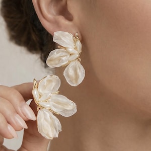 Op de afbeelding: Close-up van elegante bloemen oorbellen. De oorbellen hebben een goudkleurige zetting en delicate, gelaagde witte stoffen bloemblaadjes. Parelachtige accenten zorgen voor een verfijnd ontwerp. Ideaal voor speciale gelegenheden.