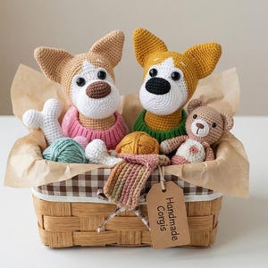Op de afbeelding: Een gevlochten mand met twee gehaakte corgi-figuren, één in een roze trui en één in een groene trui, samen met een kleine gehaakte teddybeer, bollen wol en een botvormig speeltje. Een bruin label bevat de tekst Handmade Corgis.