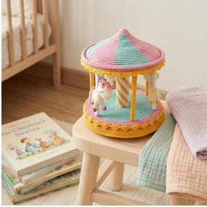May include: A handmade crochet carousel toy with a pink and teal striped roof, yellow trim, and a teal base. The carousel features white and yellow striped poles and small unicorn figures. The toy sits on a small wooden stool.