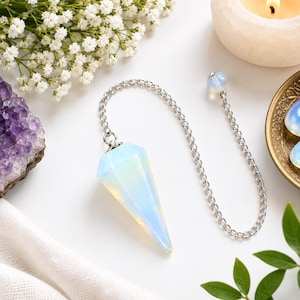 Könnte beinhalten: Ein facettiertes, durchscheinendes, hellblaues Opalith-Pendel an einer silberfarbenen Metallkette. Die Kette hat am Ende eine kleine, runde Opalith-Perle. Das Pendel hat eine spitz zulaufende, hexagonale Form und zeigt irisierende Lichtreflexe.