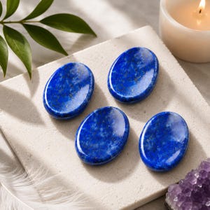May include: Four polished, oval-shaped lapis lazuli stones with a deep blue color and characteristic gold-colored pyrite flecks. The stones have a smooth, reflective surface and are arranged in a cluster on a light-colored, textured stone slab.