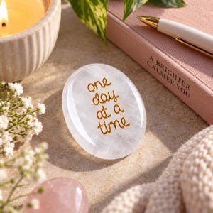 May include: A smooth, oval-shaped clear stone featuring the gold-coloured text "one day at a time" printed on its surface. The stone is positioned near a pink notebook, a white pen, a candle, and small white flowers.