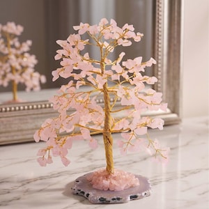 May include: A decorative tree sculpture with a gold-coloured wire trunk and branches, adorned with pink gemstone leaves. The tree sits on a grey and pink stone base. A similar tree is reflected in a mirror in the background.