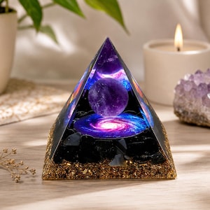 Può includere: Una piramide in resina trasparente contenente una sfera viola, una grafica di una galassia a spirale blu e rosa brillante e uno strato di base di pietre nere e trucioli di metallo dorato. La piramide ha una punta liscia e una base quadrata.