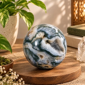 Pode incluir: Uma pedra de ágata musgo polida em forma de esfera com padrões de redemoinho em verde escuro, branco e cinza translúcido, com uma cavidade de geodo central preenchida com pequenos cristais de quartzo branco brilhantes sobre base de madeira.