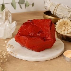 May include: A raw, unpolished chunk of vibrant red jasper stone with a jagged, irregular surface texture. The rock displays deep crimson hues and natural facets, resting on a smooth, circular white marble slab as a decorative display piece.