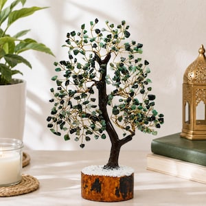Puede incluir: Una escultura de árbol decorativa con un tronco marrón oscuro y ramas hechas de alambre dorado trenzado. Las ramas están adornadas con numerosas pequeñas piedras preciosas verdes pulidas. El árbol está montado sobre una base de madera redonda.