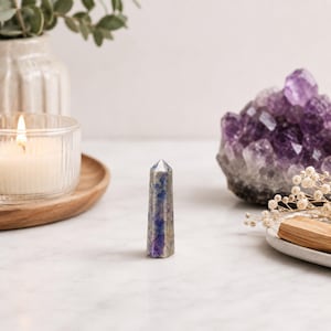 May include: A polished, six-sided crystal tower featuring a blend of deep blue, purple, and grey mineral inclusions. The stone has a pointed, faceted top and a smooth, cylindrical body, standing upright as a singular decorative mineral specimen.
