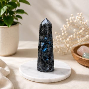 Pode incluir: Uma torre de cristal de labradorite polida com uma cor base cinzento-escura e flashes iridescentes azul-vibrantes em toda a sua superfície facetada. A pedra tem a forma de um obelisco alto de seis lados com um topo pontiagudo, sobre uma base circular de mármore branco.