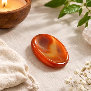 Può includere: Una pietra da meditazione in corniola di forma ovale, liscia e con una finitura lucida. La pietra presenta una sfumatura di toni arancione intenso, marrone rossastro e ambra traslucida, con motivi naturali e una rientranza concava centrale.