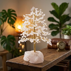 May include: A crystal tree ornament featuring clear, leaf-shaped crystals on gold wire branches. The trunk is twisted gold wire, set on a pink quartz base. The tree is displayed on a wooden surface, with a clock and books in the background.