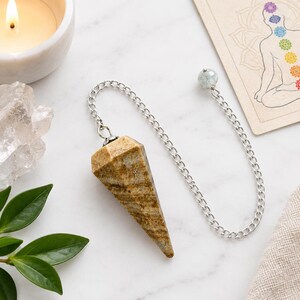 May include: A faceted, cone-shaped pendulum made of tan and brown banded stone. The pendulum is attached to a silver-toned metal chain that features a small, round, pale blue bead at the end, designed for holding or balancing during use.