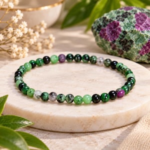 Pode incluir: Uma pulseira circular de contas com pequenas pedras esféricas polidas em tons variados de verde floresta, preto e magenta. As contas estão enfiadas num laço contínuo, exibindo padrões minerais naturais e superfícies lisas e reflexivas.