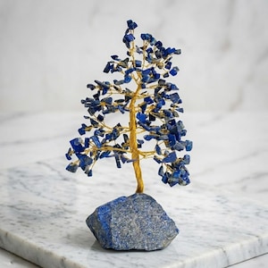 Könnte beinhalten: Eine dekorative Baumskulptur mit einem goldfarbenen Drahtstamm und Ästen. Die Blätter bestehen aus kleinen, dunkelblauen Lapislazuli-Steinen. Der Baum ist auf einem blauen Steinsockel montiert und schafft ein einzigartiges und künstlerisches Stück.