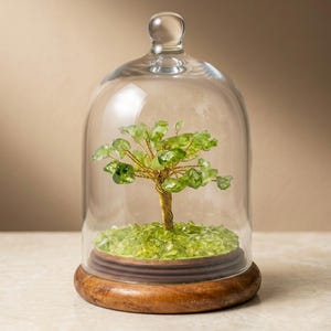 Könnte beinhalten: Dekorative Glasglocke mit einer Miniatur-Baumskulptur. Der Baum hat einen goldfarbenen Drahtstamm und Äste mit grünen Kristallblättern. Er steht auf einem Holzsockel mit einem Bett aus grünen Kristallen. Höhe ca. 20 cm.