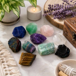 Op de afbeelding: Een verzameling van elf ruwe, ongepolijste edelstenen in diverse kleuren, waaronder diepblauw, felgroen, lichtroze, paars, transparant, bruin gestreept en zwart. De stenen liggen op een witte ondergrond naast een klein houten doosje.