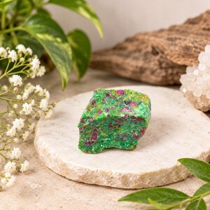 May include: A raw, unpolished mineral specimen featuring a vibrant green matrix speckled with deep magenta and pink crystalline inclusions. The stone has a rough, jagged texture and sits on a circular, light-coloured stone coaster.