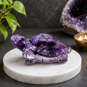 May include: A carved turtle figurine crafted from deep purple amethyst crystal. The sculpture features a detailed shell with geometric facets, a smooth head, and four legs, showcasing the natural translucent and opaque variations of the stone.