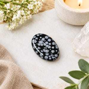 Pode incluir: Uma pedra preciosa de obsidiana floco de neve de formato oval, com uma superfície preta lisa e polida e distintos padrões radiais brancos em forma de estrela espalhados pela pedra, criando um contraste natural entre a base escura e as inclusões claras.
