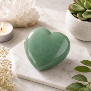 Può includere: Una pietra verde liscia e lucida scolpita a forma di cuore. Il cuore presenta una superficie brillante e riflettente con sottili variazioni nelle tonalità verde chiaro e scuro, mostrando una forma solida e arrotondata con una fessura in alto.