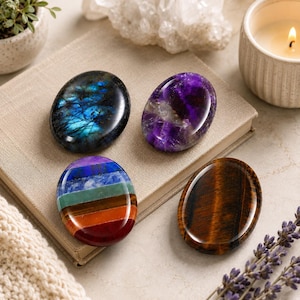 May include: Four smooth, oval-shaped polished stones resting on a closed book. One stone is dark blue with iridescent flashes, one is deep purple, one features horizontal stripes of seven colors, and one is brown with a chatoyant banded pattern.