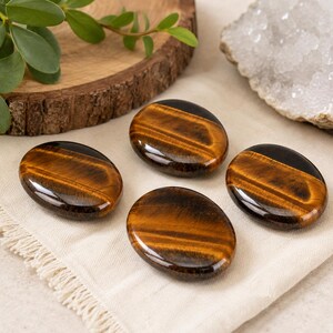 Puede incluir: Cuatro piedras de ojo de tigre pulidas de forma ovalada con un acabado suave y brillante. Cada piedra muestra bandas chatoyantes características de color marrón dorado y marrón oscuro que crean un aspecto brillante y estratificado.