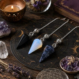May include: Three pointed crystal pendulums attached to silver-toned metal chains. One pendulum is solid black, one is translucent white with an iridescent glow, and one is a mottled dark blue. Each chain features a decorative silver bead.