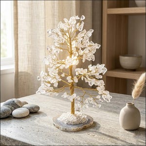 Könnte beinhalten: Eine dekorative Kristallbaumskulptur mit einem goldfarbenen Drahtstamm und Ästen. Die Blätter des Baumes bestehen aus klaren Kristallen. Der Baum steht auf einem grau-weißen Achatfuß. Die Skulptur wird auf einer hellgrauen Oberfläche präsentiert.