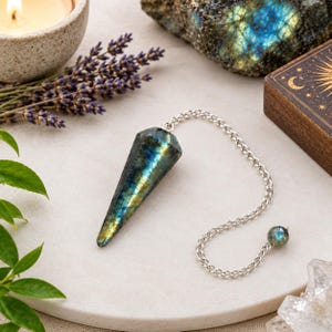 Könnte beinhalten: Ein spitz zulaufendes, facettiertes Labradorit-Pendel mit irisierenden blauen und goldenen Reflexen, befestigt an einer silberfarbenen Metallkette. Die Kette endet in einer kleinen, runden Labradorit-Perle, die für ein ausgewogenes Gewicht sorgt.