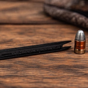 May include: A black, slotted tool and a bullet with a silver tip, copper midsection, and brass base rest on a wooden surface. The tool is long and thin with a forked end. A patterned fabric is in the background.