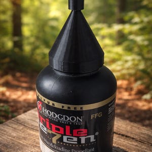 May include: A black bottle of Hodgdon Triple Seven muzzleloading propellant. The bottle has a black conical cap and gold lettering. The label reads "Triple Seven" in red and white text. The background is a blurred forest.