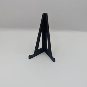 May include: A black display stand with a triangular shape. The stand has a flat base with two small feet and a vertical support that tapers to a point. The stand is designed to hold a small object.