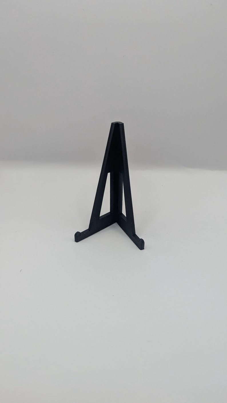 May include: A black display stand with a triangular shape. The stand has a flat base with two small feet and a vertical support that tapers to a point. The stand is designed to hold a small object.
