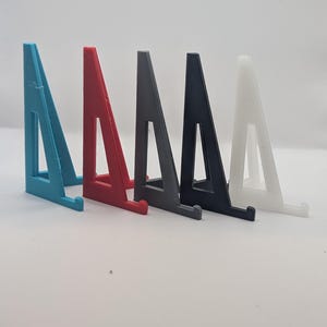 May include: A row of five 3D-printed display stands in various colors. The stands are shaped like triangles with a small ledge at the bottom to hold an item. Colors include teal, red, gray, black, and white.