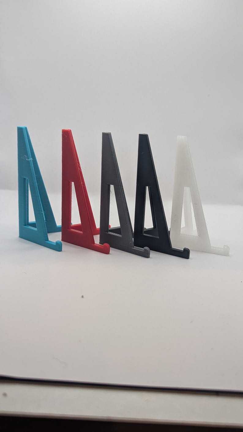 May include: A row of five 3D-printed display stands in various colors. The stands are shaped like triangles with a small ledge at the bottom to hold an item. Colors include teal, red, gray, black, and white.