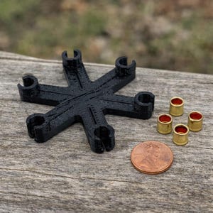 May include: A black, star-shaped object with six curved arms, alongside five small, gold-colored cylinders with red interiors. A penny is included for size comparison. The items are arranged on a weathered wooden surface.