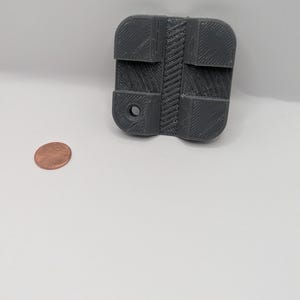 May include: A gray 3D-printed object with a rectangular shape and a textured surface. The object has a central patterned design and a circular hole. A copper penny is next to the object.