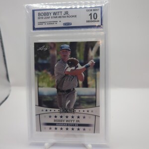 May include: A baseball card featuring Bobby Witt Jr. in a Kansas City Royals uniform. The card is graded GEM MINT 10 and is a 2019 Leaf Star #ST04 Rookie card. The card is encased in a protective holder.