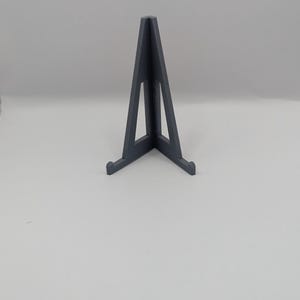 May include: A dark gray, triangular display stand with a wide base and a central support. The stand is made of a solid material and is designed to hold items upright. The design is simple and modern, suitable for various display purposes.