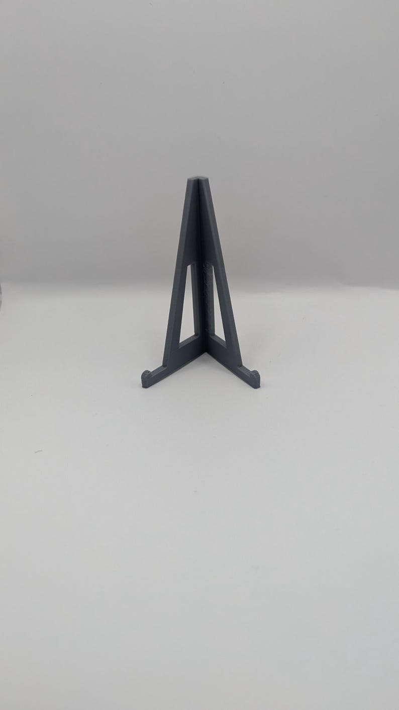 May include: A dark gray, triangular display stand with a wide base and a central support. The stand is made of a solid material and is designed to hold items upright. The design is simple and modern, suitable for various display purposes.