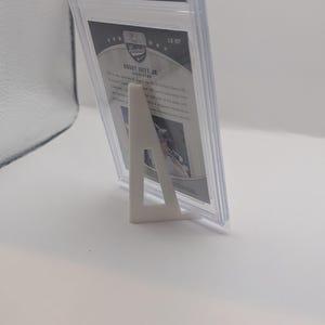 May include: A graded trading card in a clear plastic case, supported by a white stand. The card features text and a logo from CCG grading. The card is angled, showcasing the details of the card and the protective case.