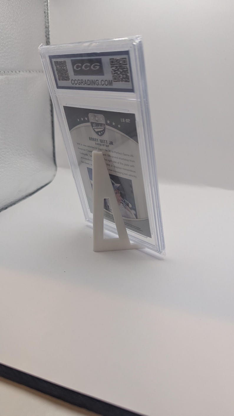 May include: A graded trading card in a clear plastic case, supported by a white stand. The card features text and a logo from CCG grading. The card is angled, showcasing the details of the card and the protective case.