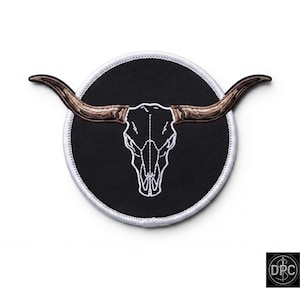 Può includere: Una toppa ricamata circolare con sfondo nero e il contorno bianco cucito di un teschio di longhorn. Il teschio di longhorn presenta corna marroni testurizzate che si estendono oltre il bordo circolare della toppa.