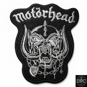 May include: A black embroidered patch featuring the white text "motörhead" at the top. Below the text is a detailed white stitched graphic of a snarling creature with large horns, a helmet, and a chain necklace with a small cross and skull charm.