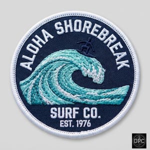 May include: A circular embroidered patch with a dark blue background featuring a light blue and white ocean wave graphic, a small surfer silhouette, and the white text ALOHA SHOREBREAK SURF CO. EST. 1976, all enclosed by a white stitched border.